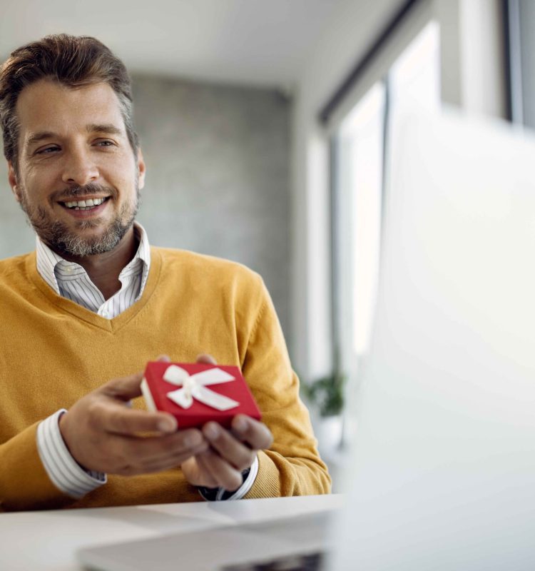 happy-businessman-holding-small-gift-box-while-making-video-call-laptop